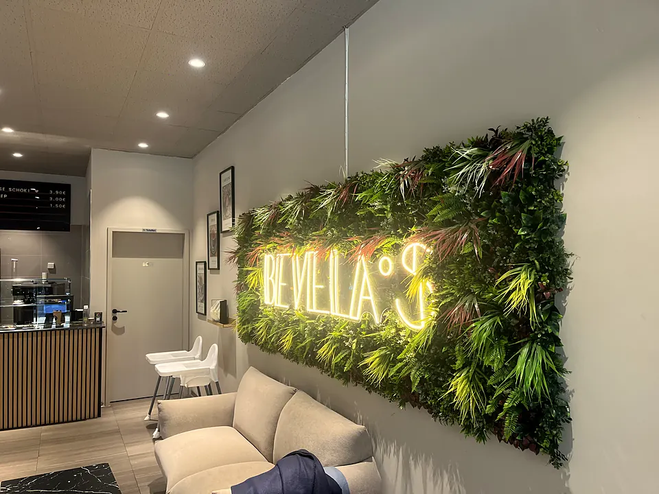 Sitzecke mit heller Couch unter der Pflanzenwand mit dem Neon-Schriftzug Bevela's